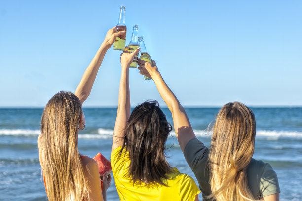 三个快乐的女孩举杯庆祝暑假，看着户外的地平线海滩。不知名的朋友在日落时分的海边度假。快乐，自由，genz和政党的概念图片下载