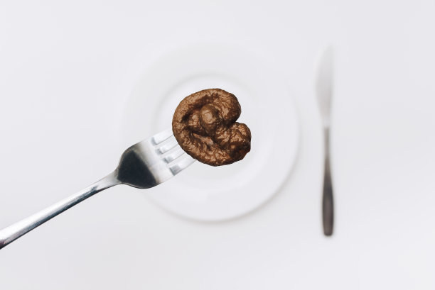 一只猫的人造塑料粪便躺在叉子上，旁边是白色的盘子和刀。假新闻的概念，蹩脚的厨师，蹩脚的餐厅和劣质的食物。糟糕的笑话，屎的味道。图片下载