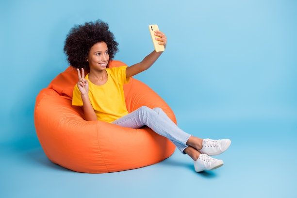 小黑皮肤棕色头发的小女孩穿休闲服装拿着手机使自拍v形标志孤立在蓝色背景图片下载