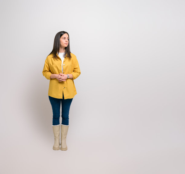沉思的年轻女子穿着衬衫和橡胶靴，站在背景中思考。沉思的美丽女性，双手紧握，看着别处，自信地摆姿势图片下载