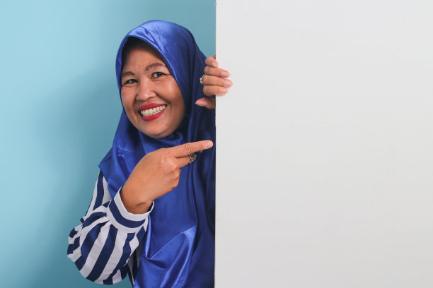 兴奋的亚洲妇女戴着头巾，指着空白的白色横幅招牌，上面写着文案。蓝色背景。图片下载