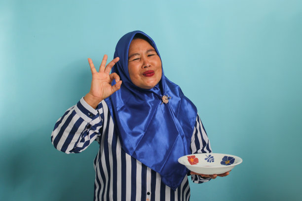 快乐的亚洲女人，中年，蓝色头巾，用空盘子亲吻手指，蓝色背景。图片下载