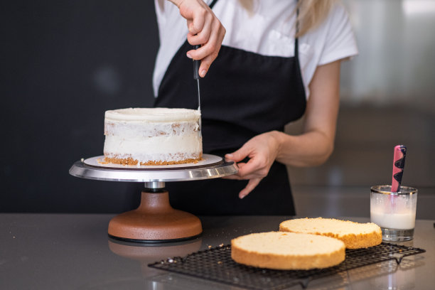 穿着黑色围裙的家庭主妇在厨房用新鲜的白奶油涂抹有机蛋糕层图片下载