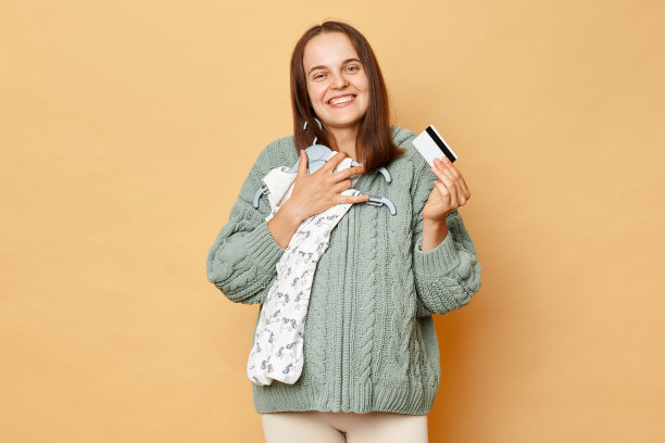 快乐的微笑，美丽的未来母亲，年轻的孕妇，婴儿紧身衣被隔离在米色背景上，拥抱着孩子的衣服，微笑着对着镜头期待着孩子。图片下载