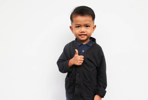 一个微笑的蹒跚学步的男孩看着镜头，举起双手竖起大拇指，孤立在白色的背景上图片下载