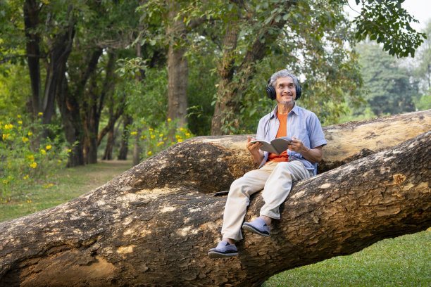快乐的老男人坐在夏日公园的树干上，听着音乐，读着书，宁静的老男人在大自然中放松图片下载