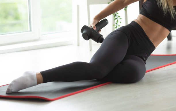 年轻健康的妇女练习自我按摩技术的臀部应用治疗性的冲击按摩枪坐在家里的瑜伽垫。图片下载