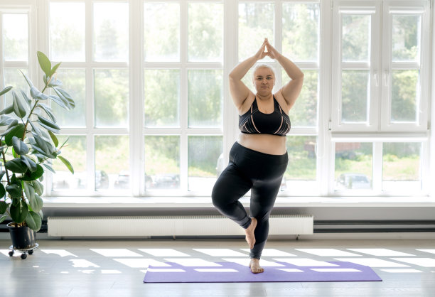 胖乎乎的女人站在树上做瑜伽。女性通过站在运动垫子上健身来保持平衡。房间里有大而高的窗户，让阳光充足图片下载
