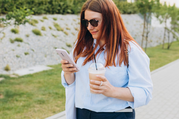 户外生活时尚肖像的女人摆姿势在街上喝冰沙或冰咖啡。一位女士手持环保或可重复使用的购物袋，使用智能手机图片下载