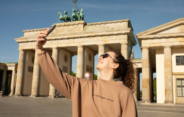 在柏林旅行。在德国柏林的勃兰登堡门前，一名开心的女游客用手机自拍。图片下载