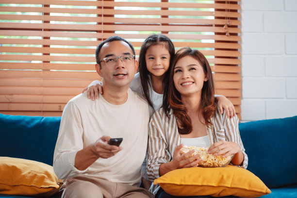 亚洲家庭由父母和女儿在家里客厅的沙发上看电视或电影，吃爆米花组成。享受放松的快乐。图片下载