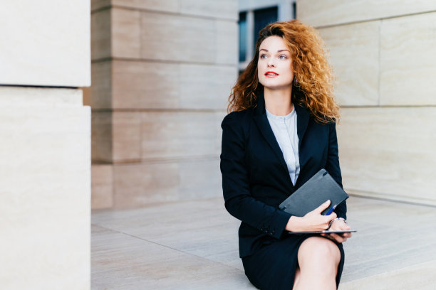 漂亮的女商人:华丽的头发，蓝眼睛，红唇。身穿黑色西装，手持袖珍书和平板电脑。把沉思的目光放到一边。图片下载