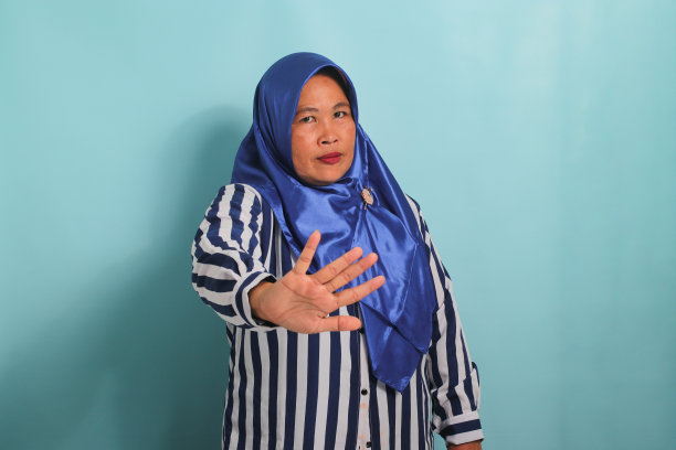 严肃的亚洲妇女戴着蓝色头巾，穿着条纹衬衫，在蓝色背景上做出停止或拒绝的手势图片下载