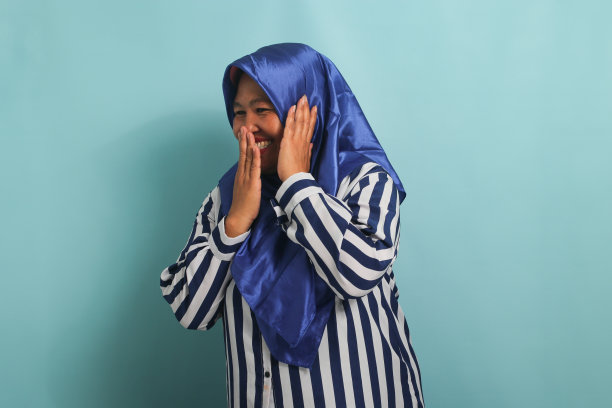 好奇的亚洲妇女戴着蓝色头巾，对八卦大笑，手掌挨着耳朵，孤立在蓝色背景下图片下载