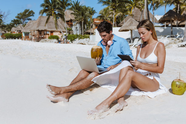 西班牙年轻的数字游牧民夫妇坐在海滩上，用笔记本电脑和平板电脑享受旅行冒险假期，在墨西哥、拉丁美洲、加勒比海和热带目的地工作图片下载