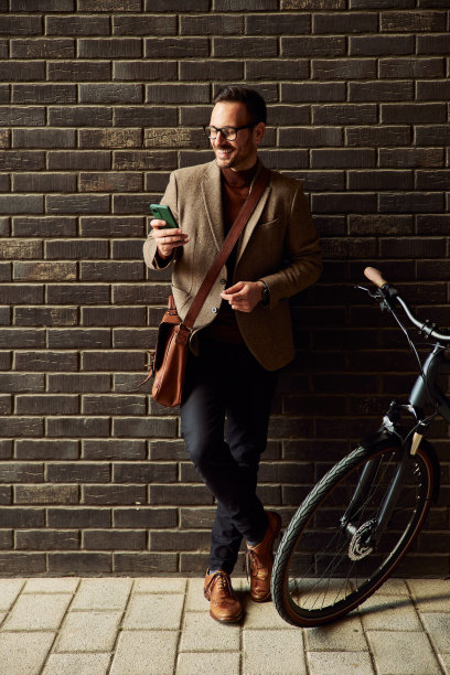 一名商人骑着自行车，靠在砖墙上使用手机。图片下载