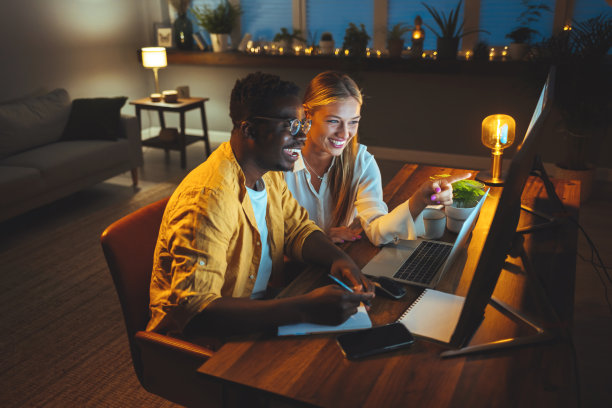 英俊的男人晚上坐在电脑前工作，他的妻子坐在他旁边。图片下载