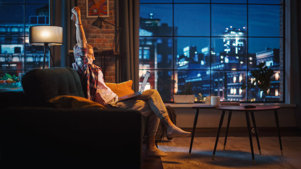 年轻美丽的女人坐在沙发上，使用笔记本电脑在晚上的时尚阁楼公寓。创意女性庆祝成功的消息。从大窗户看城市。图片下载