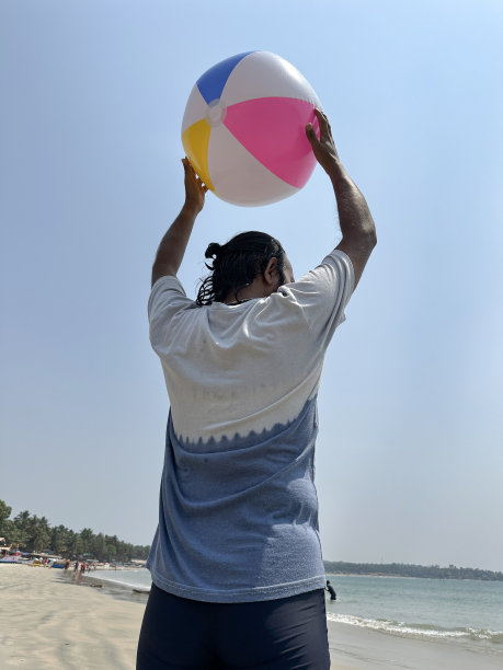 印度男子身穿冲浪短裤和t恤，站在海边的沙滩上，头顶沙滩球，色彩鲜艳的沙滩玩具，船只，有棕榈树的林地，清澈的蓝色阳光明媚的天空，后景图片下载