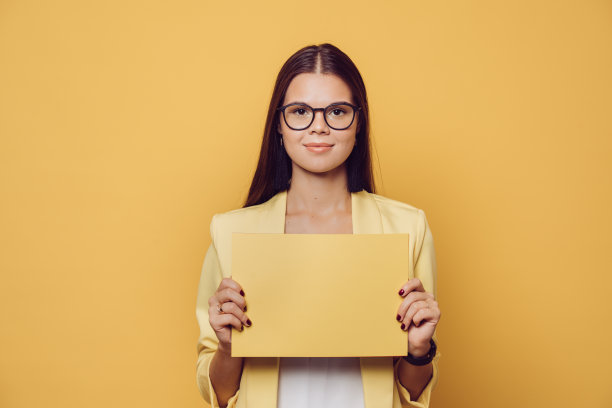 年轻自信的黑发女子戴着眼镜，长发穿着黄色夹克，在黄色的工作室背景上拿着黄色的纸。图片下载