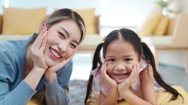 美丽年轻的亚洲母亲和小女儿躺在客厅的地板上，双手放在脸颊上，对着镜头微笑。幸福家庭，单亲妈妈，母子关系概念图片下载