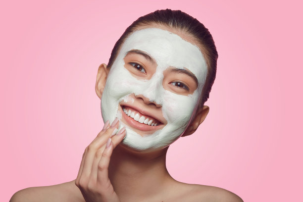 微笑着的亚洲女人戴着口罩，健康美丽的女性在脸上涂着绿色的粘土面膜，站在粉红色的隔离上。图片下载