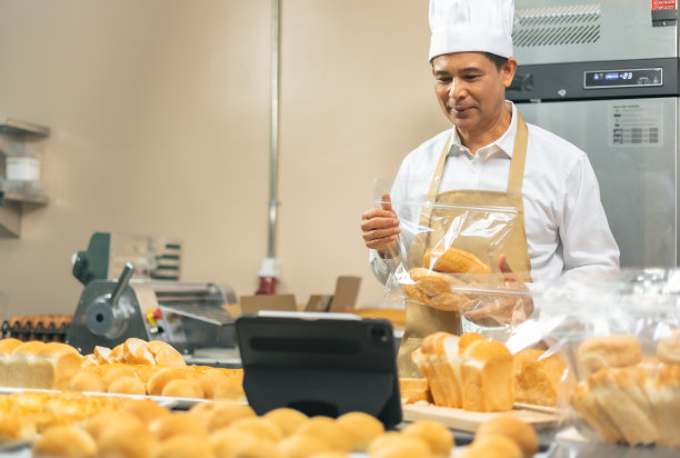 穿着围裙的资深亚洲男厨师，通过移动应用技术直播小企业博主，烘焙销售在线推广，新鲜面包。图片下载