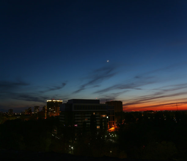 一个年轻的月亮在深蓝色的天空下成长的彩色照片。日落时美丽的城市夜景。天际线的房屋，结构和建筑物。图片下载