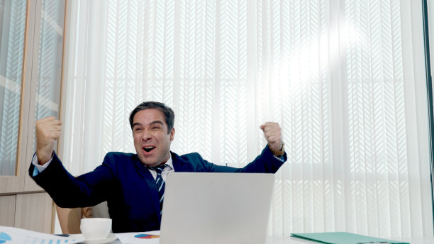 快乐的商人兴奋惊讶的工作在白色的办公桌与pc笔记本电脑扔纸帐户文件图片下载