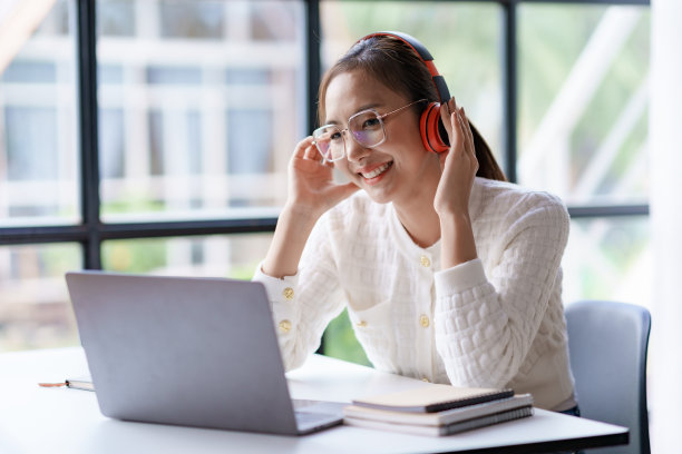 可爱的年轻亚洲女子戴着耳机，愉快地在笔记本电脑上听音乐，在工作休息时享受工作音乐。图片下载