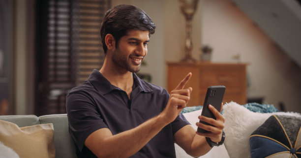 快乐的年轻印度男子在家舒适地使用他的智能手机，英俊，微笑和积极。他正在浏览社交媒体或从事虚拟远程工作。图片下载