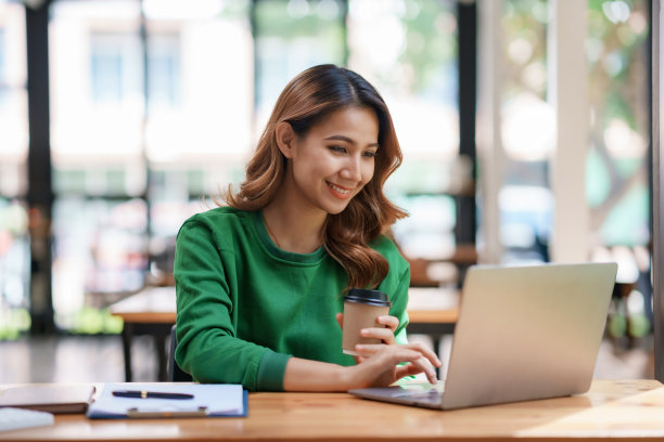 漂亮的亚洲女商人愉快地坐在她的笔记本电脑上工作，喝着咖啡，在办公室里笑得很开心。图片下载