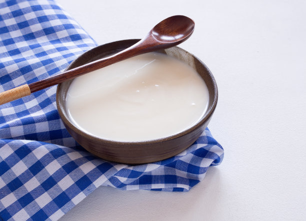 自制的希腊酸奶或酸奶油，装在旧陶碗里，配上木勺和白色木制背景上的蓝色方格织物或餐巾。图片下载