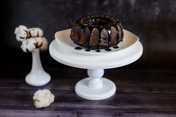 白色盘子里放着巧克力蛋糕，上面有糖霜，架子上有一个黑色背景的花瓶，花瓶上有一朵棉花花图片下载