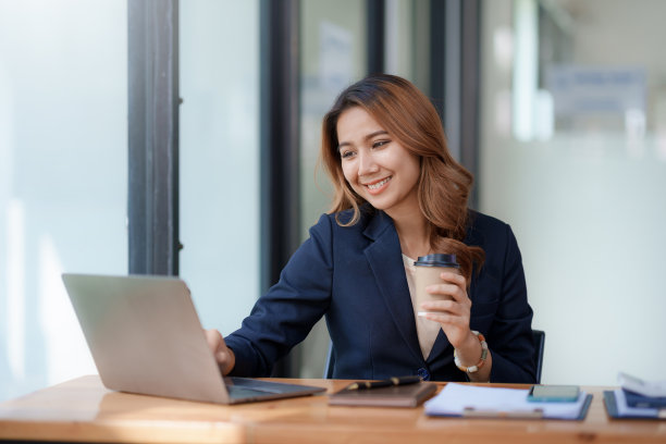 美丽的亚洲女商人坐在她的笔记本电脑上愉快地工作，微笑着喝着咖啡，看着镜头，在办公室里看起来很明亮。图片下载