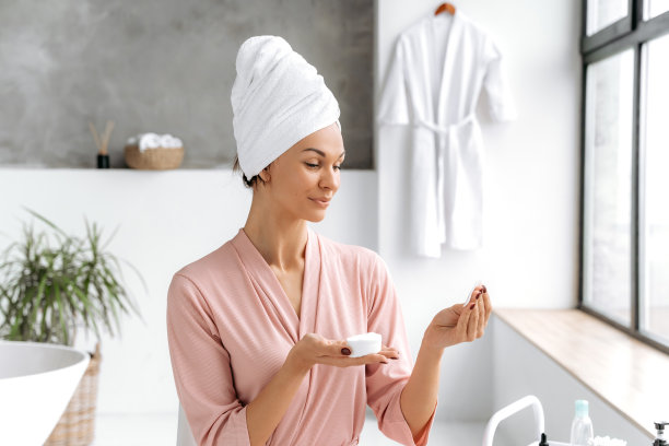 漂亮的白人女人穿着浴袍，头上缠着毛巾，拿着白色的棉垫清洁她敏感的皮肤，坐在浴室里使用护肤品。皮肤清洁图片下载