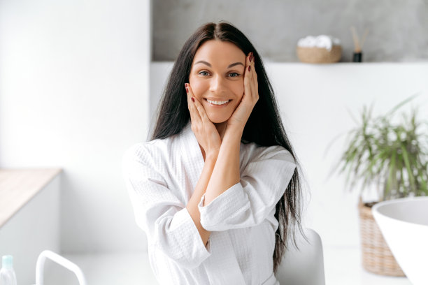 照片中，一位漂亮、整洁的白人年轻女子，留着长长的黑发，穿着浴袍，站在浴室里，双手贴近脸，看着镜头，开心地笑着图片下载