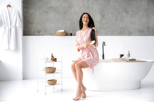 一个可爱快乐的黑发白人长头发女人的全身照片，穿着晨衣，坐在浴室里洗澡，做着例行的女性护理程序，梳理她的头发，微笑着图片下载