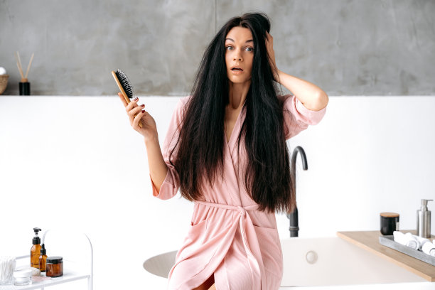 拿着梳子的女人。震惊而焦虑的白人妇女穿着浴袍，一头浓密的黑色长发，坐在浴室里，手里拿着一把梳子，梳头后忧心忡忡地看着镜头图片下载