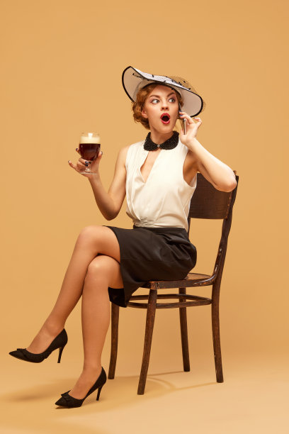 漂亮的女人穿着优雅的衣服，戴着帽子，拿着一杯啤酒，在米色的背景下用手机说话。八卦,闲谈图片下载