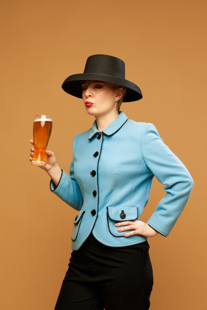 一个穿着优雅服装的漂亮女人，在米色背景上拿着一杯泡沫啤酒。黑帽子女士图片下载