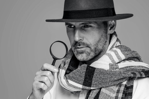 男侦探带着放大镜，戴着时尚的围巾和棕色帽子，在工作室背景上摆姿势。图片下载