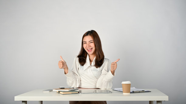一位兴高采烈的亚洲女商人坐在她的办公桌前，竖起大拇指。图片下载