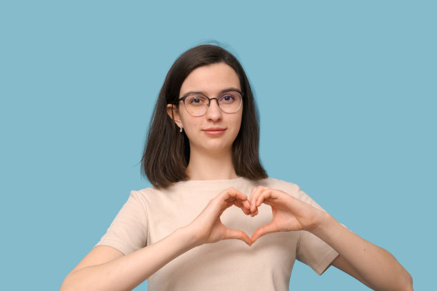 漂亮的女学生戴着眼镜，用手做了一个心形，在蓝色的背景上微笑。爱,关系图片下载