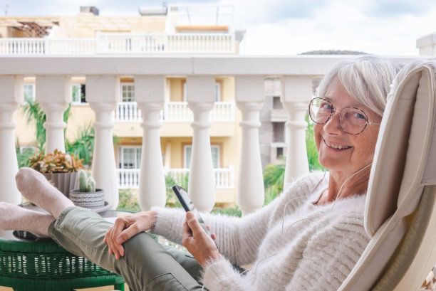 快乐的年长妇女放松在家里阳台上拿着手机-成年人有吸引力的女性享受安静的生活方式，户外听音乐图片下载