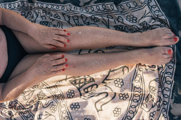 度假的退休老妇人坐在海边的毛巾上。俯拍的照片上，一位面目全非的老妇人坐在曼荼罗沙滩浴巾上，伸开双腿，双手放在膝盖上。图片下载