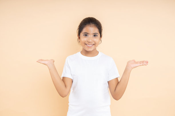 快乐的印度小女孩穿着白色t恤，张开双臂，孤立在米色的工作室背景上，看这里，放上你的广告和促销。图片下载