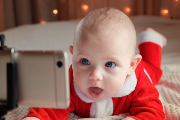 微笑的宝宝穿着红色圣诞老人服装，看着手机进行视频通话。祝贺你。可爱的孩子用智能手机交流和玩得开心。圣诞快乐，新年快乐。图片下载