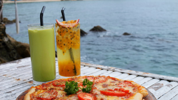 两种异国情调的鸡尾酒和新鲜出炉的披萨在海滨酒店咖啡馆的慢动作镜头。绿松石海与光波的背景。宁静祥和的休息和放松的地方。图片下载