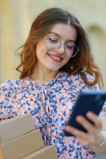 一个快乐的女人，穿着裙子，戴着眼镜，手里拿着纸板箱，手里拿着装有邮件应用程序的智能手机。图片下载